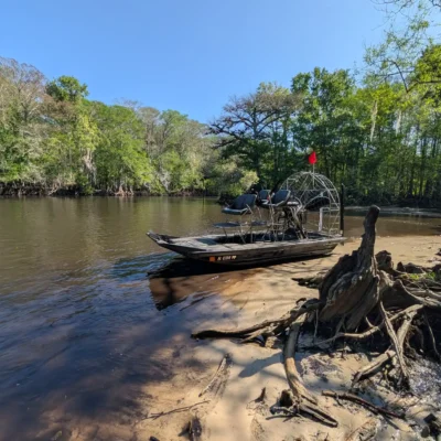 An airboat with a red flag is partially beached on a sandy bank beside a calm river, ready for an exciting excursion, surrounded by trees under a clear blue sky.