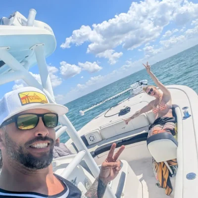 Two people on a boat smiling and making peace signs during an exciting excursion, with blue skies, clouds, and ocean water in the background.