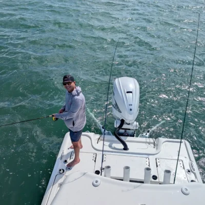 A person stands barefoot at the back of a white boat, enjoying a fishing excursion in open water on a sunny day.
