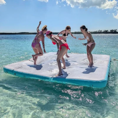 Four women in swimsuits enjoy an excursion, balancing and playing on an inflatable floating mat in clear, shallow water under a sunny sky with some clouds.