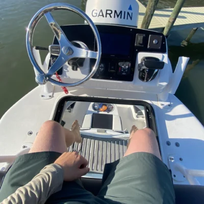 Person sitting at the helm of a boat, ready for an exciting excursion, with Garmin navigation equipment, wearing green shorts and a beige shirt, hand on their lap and feet resting on the deck.