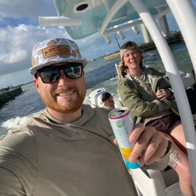 A man wearing sunglasses and a cap holds a canned drink and smiles at the camera during an excursion, sitting on a boat with two other people on a sunny day.