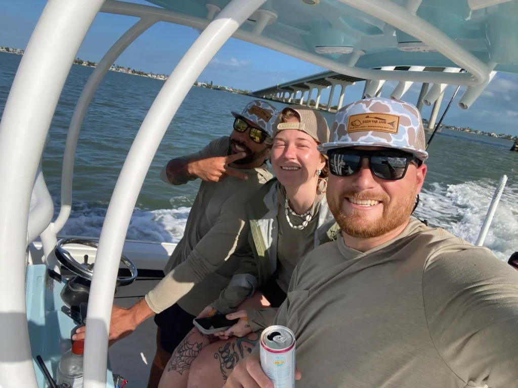 Three people on a boat excursion smiling, wearing sunglasses and hats, with one holding a drink; ocean and sky visible in the background.