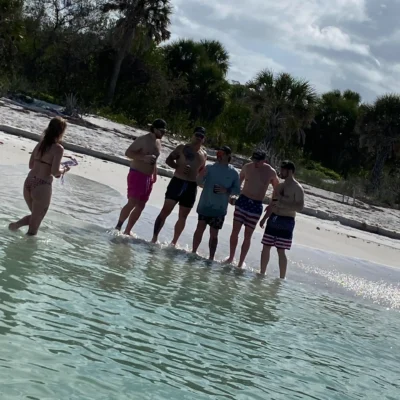 Six men in swimwear pose for a photo on a sandy beach during an excursion, while a woman in a bikini stands in shallow, clear water holding a phone with trees visible in the background.