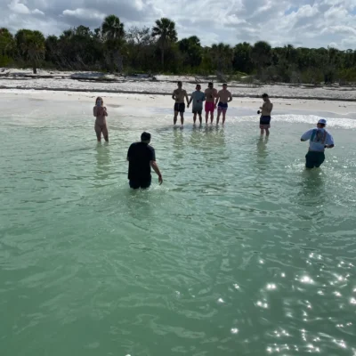 A group of people enjoys an excursion, standing in shallow, clear water near a sandy beach with trees in the background under a partly cloudy sky.