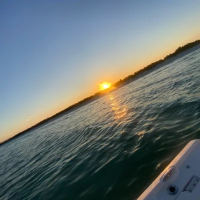 Sunset over a calm body of water with gentle waves, the shoreline in the distance, and the edge of a boat visible in the foreground—perfect for an evening excursion.