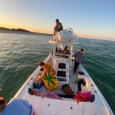 A group of people are relaxing on a motorboat during an evening excursion at sunset, with one person standing on the upper deck and others sitting or lying on the main deck.